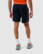 A person wearing a bright orange shirt, navy blue Psycho Bunny MENS SONTERRA STRIPE SPORT SHORT - B6R464C200 with orange stripes along the sides, white socks, and colorful sneakers with orange, blue, and white accents stands facing away from the camera. The stretch fabric of the outfit ensures comfort and mobility.