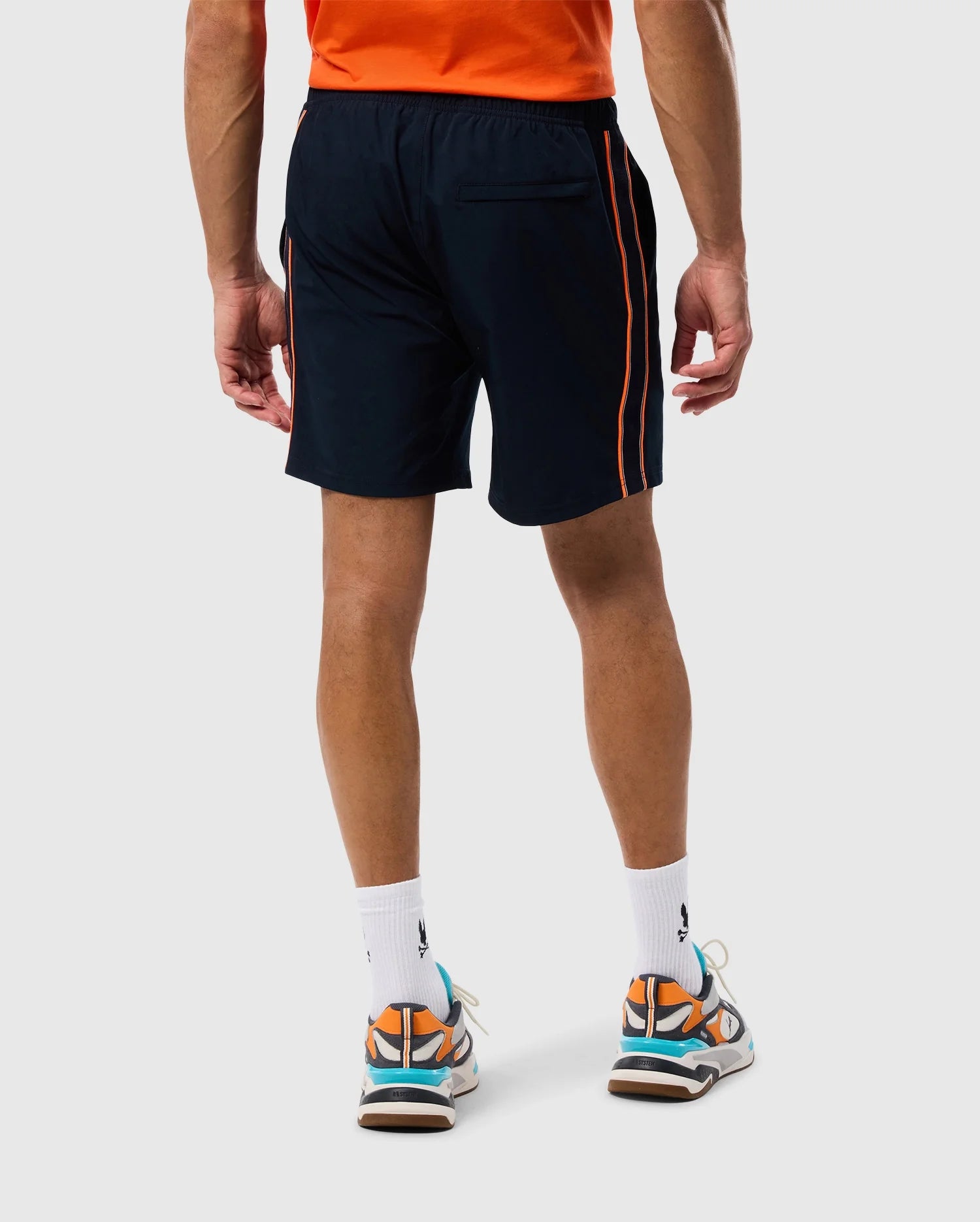 A person wearing a bright orange shirt, navy blue Psycho Bunny MENS SONTERRA STRIPE SPORT SHORT - B6R464C200 with orange stripes along the sides, white socks, and colorful sneakers with orange, blue, and white accents stands facing away from the camera. The stretch fabric of the outfit ensures comfort and mobility.