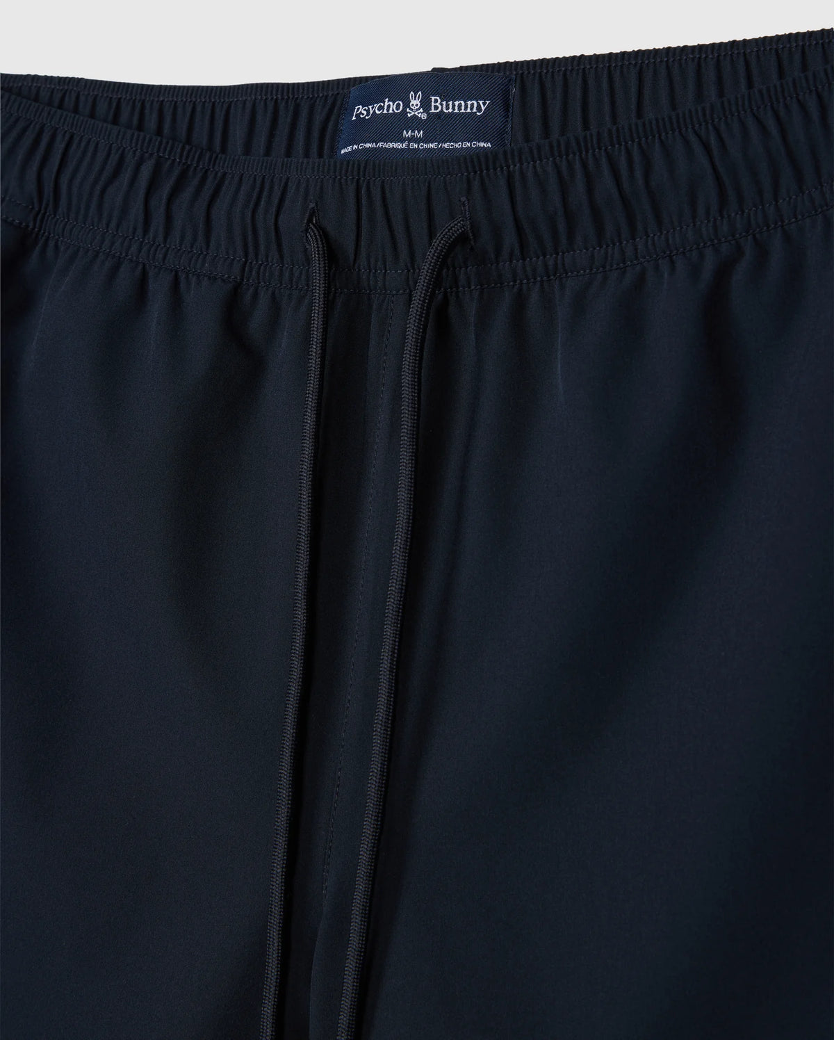 Close-up of a pair of dark blue men's sport shorts with an elastic waistband and drawstring. Made from quick-drying stretch fabric, the label reads "Psycho Bunny" at the back. These are the MENS SONTERRA STRIPE SPORT SHORT - B6R464C200 by Psycho Bunny.