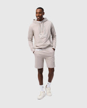A man is smiling and walking while wearing a light gray hoodie made from a high-quality cotton blend with a graphic design on the front, matching Psycho Bunny's MENS WALTER LIGHTWEIGHT SWEATSHORT (B6R477D200), white socks, and white sneakers. The regular fit hoodie features a drawstring, and his hands are in the hoodie's front pocket. The background is plain white.
