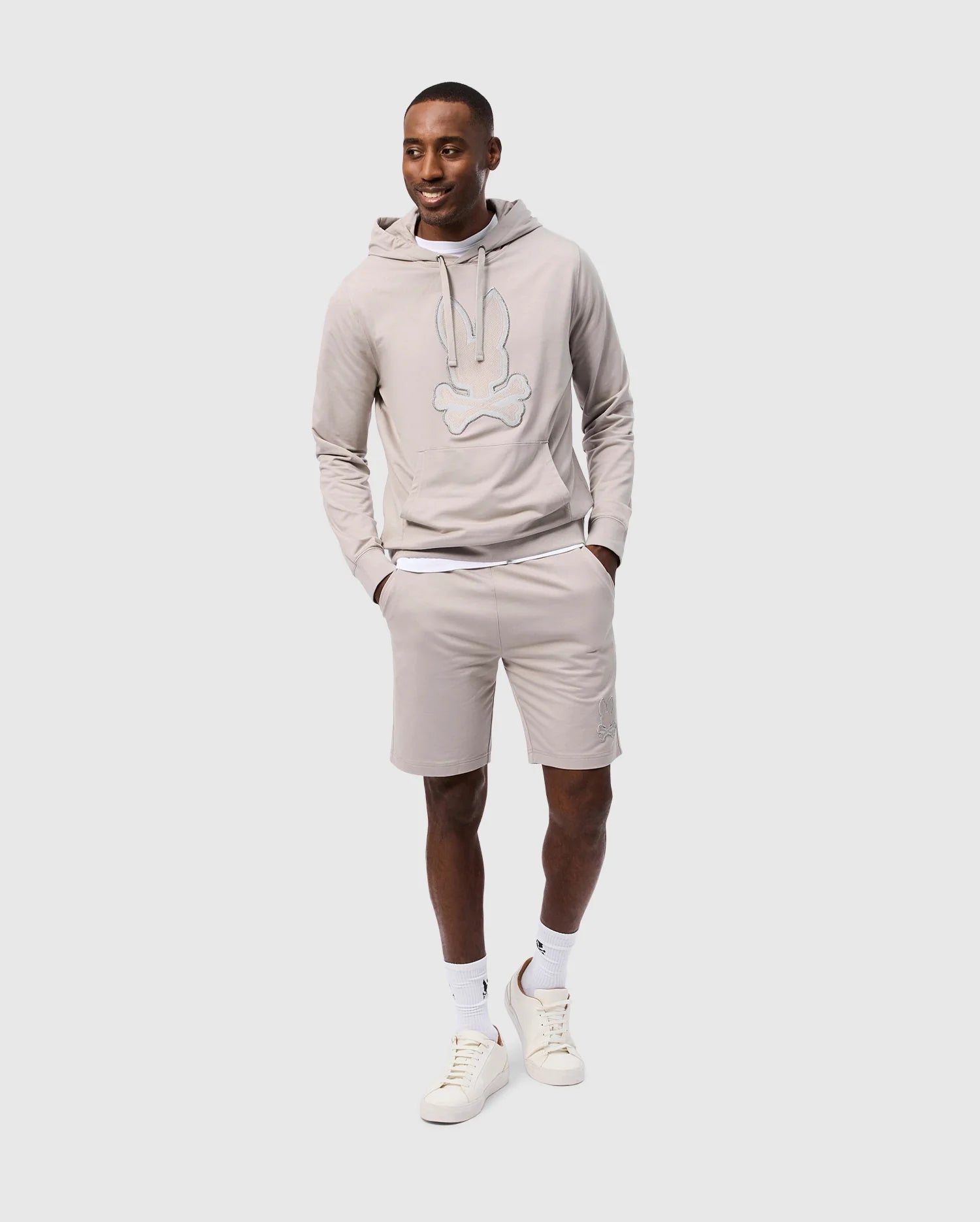 A man is smiling and walking while wearing a light gray hoodie made from a high-quality cotton blend with a graphic design on the front, matching Psycho Bunny's MENS WALTER LIGHTWEIGHT SWEATSHORT (B6R477D200), white socks, and white sneakers. The regular fit hoodie features a drawstring, and his hands are in the hoodie's front pocket. The background is plain white.