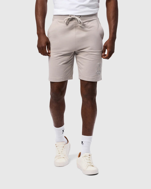 A person is wearing a high-quality cotton blend white t-shirt tucked into Psycho Bunny's MENS WALTER LIGHTWEIGHT SWEATSHORT - B6R477D200, which are regular fit light beige shorts with a drawstring at the waist. They are also wearing white crew socks with small black logos and white sneakers with beige accents. The background is plain light gray.