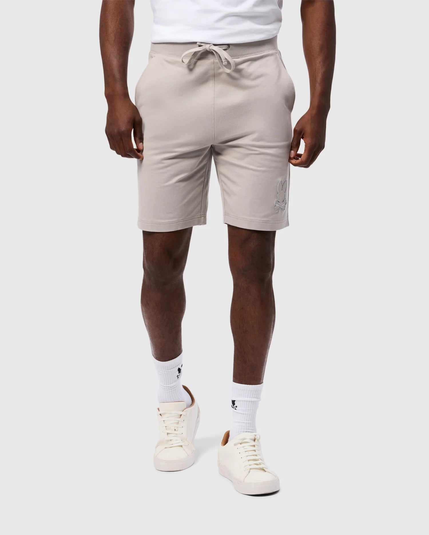 A person is wearing a high-quality cotton blend white t-shirt tucked into Psycho Bunny's MENS WALTER LIGHTWEIGHT SWEATSHORT - B6R477D200, which are regular fit light beige shorts with a drawstring at the waist. They are also wearing white crew socks with small black logos and white sneakers with beige accents. The background is plain light gray.