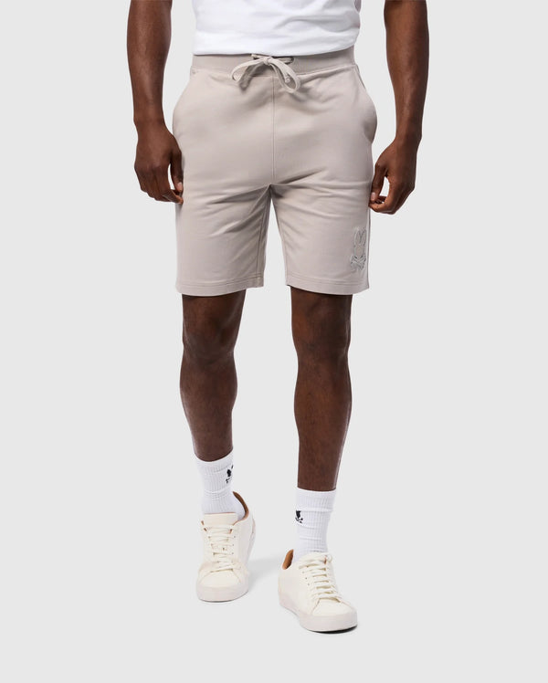 A person is wearing a high-quality cotton blend white t-shirt tucked into Psycho Bunny's MENS WALTER LIGHTWEIGHT SWEATSHORT - B6R477D200, which are regular fit light beige shorts with a drawstring at the waist. They are also wearing white crew socks with small black logos and white sneakers with beige accents. The background is plain light gray.
