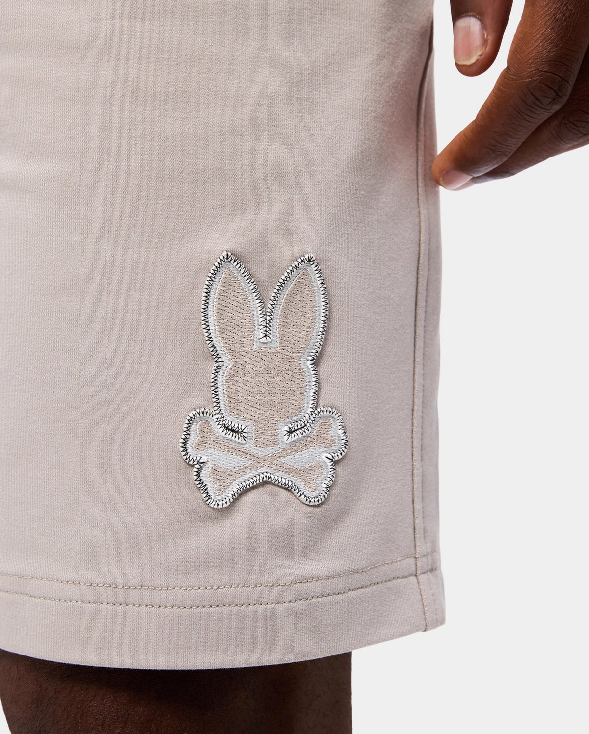 Close-up of the lower part of a person's beige, MENS WALTER LIGHTWEIGHT SWEATSHORT - B6R477D200 by Psycho Bunny, featuring an embroidered patch of a bunny head above crossbones on the left leg. Made from high-quality cotton blend, these regular fit sweatshorts offer both style and comfort. The person's hand is partially visible in the upper right corner of the image.