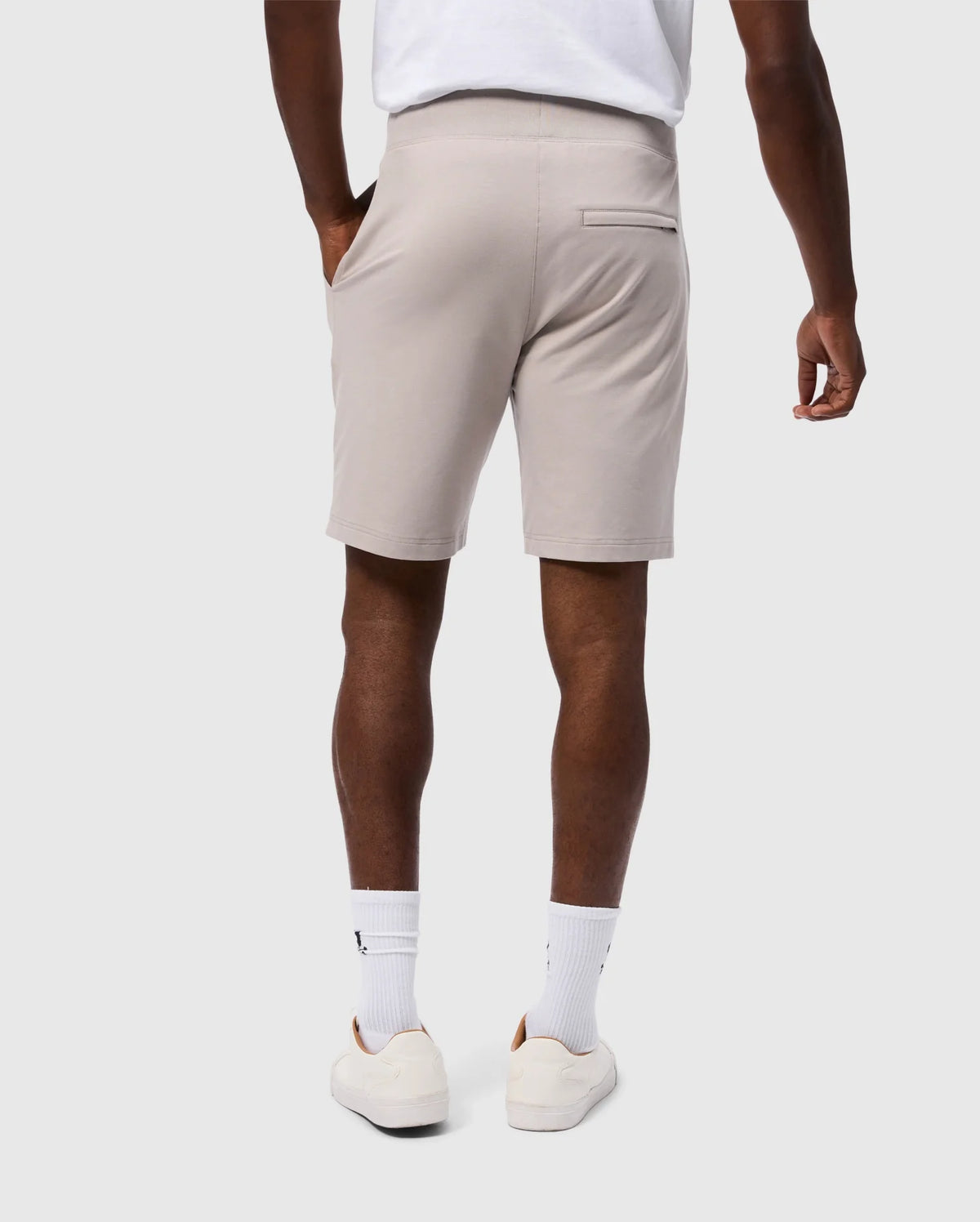 A person wearing a white T-shirt, MENS WALTER LIGHTWEIGHT SWEATSHORT - B6R477D200 by Psycho Bunny in beige, white socks, and white sneakers stands facing away. Their left hand is in their pocket, and the background is plain white.