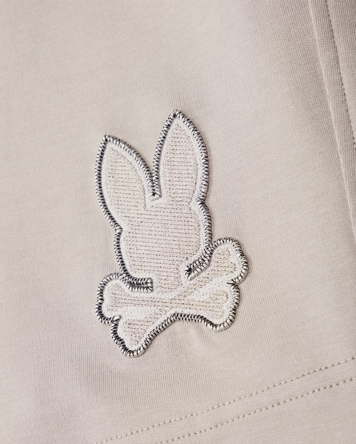A close-up of a fabric patch from the Psycho Bunny MENS WALTER LIGHTWEIGHT SWEATSHORT - B6R477D200, showcasing an embroidered design of a rabbit head and crossbones in white and grey stitching on a light beige background, crafted from high-quality cotton blend.