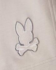 A close-up of a fabric patch from the Psycho Bunny MENS WALTER LIGHTWEIGHT SWEATSHORT - B6R477D200, showcasing an embroidered design of a rabbit head and crossbones in white and grey stitching on a light beige background, crafted from high-quality cotton blend.