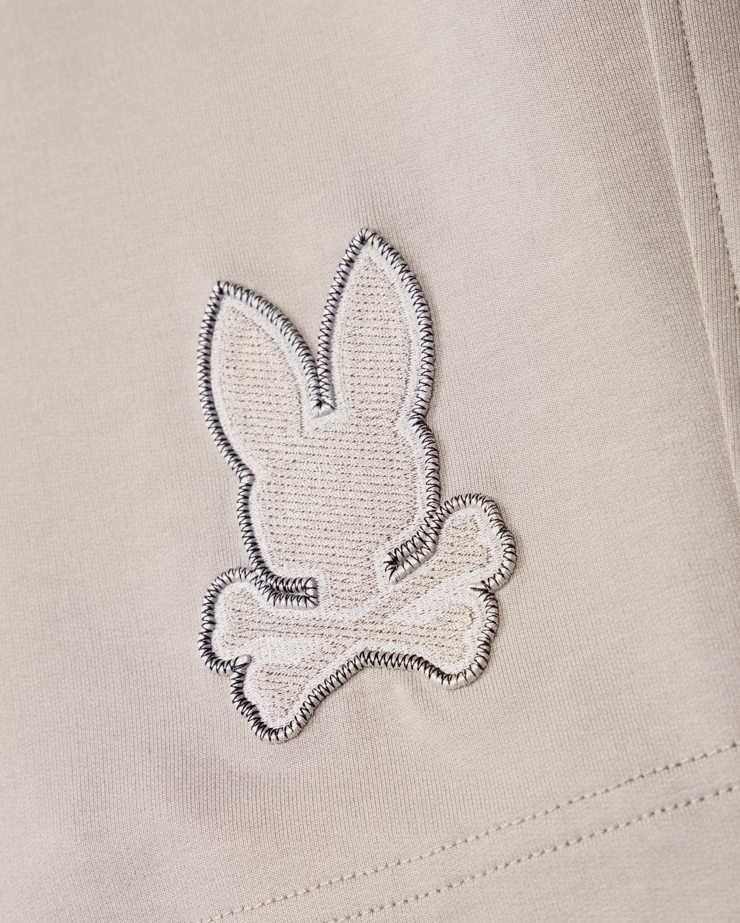 A close-up of a fabric patch from the Psycho Bunny MENS WALTER LIGHTWEIGHT SWEATSHORT - B6R477D200, showcasing an embroidered design of a rabbit head and crossbones in white and grey stitching on a light beige background, crafted from high-quality cotton blend.
