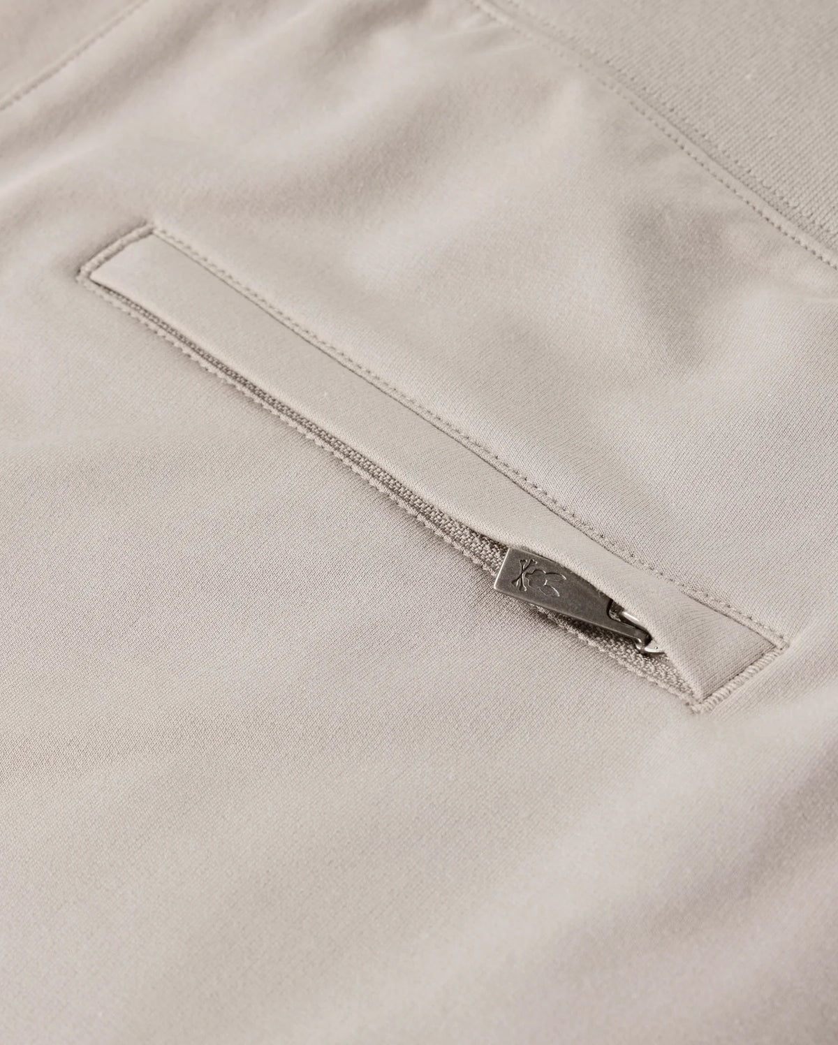 Close-up of the *MENS WALTER LIGHTWEIGHT SWEATSHORT - B6R477D200* by Psycho Bunny showcasing a light beige fabric with a horizontal zippered pocket. The zipper includes a metallic pull tab for easy access, and the surrounding fabric appears soft and smooth. This high-quality cotton blend ensures a uniform, finely woven texture in this lightweight sweatshort.