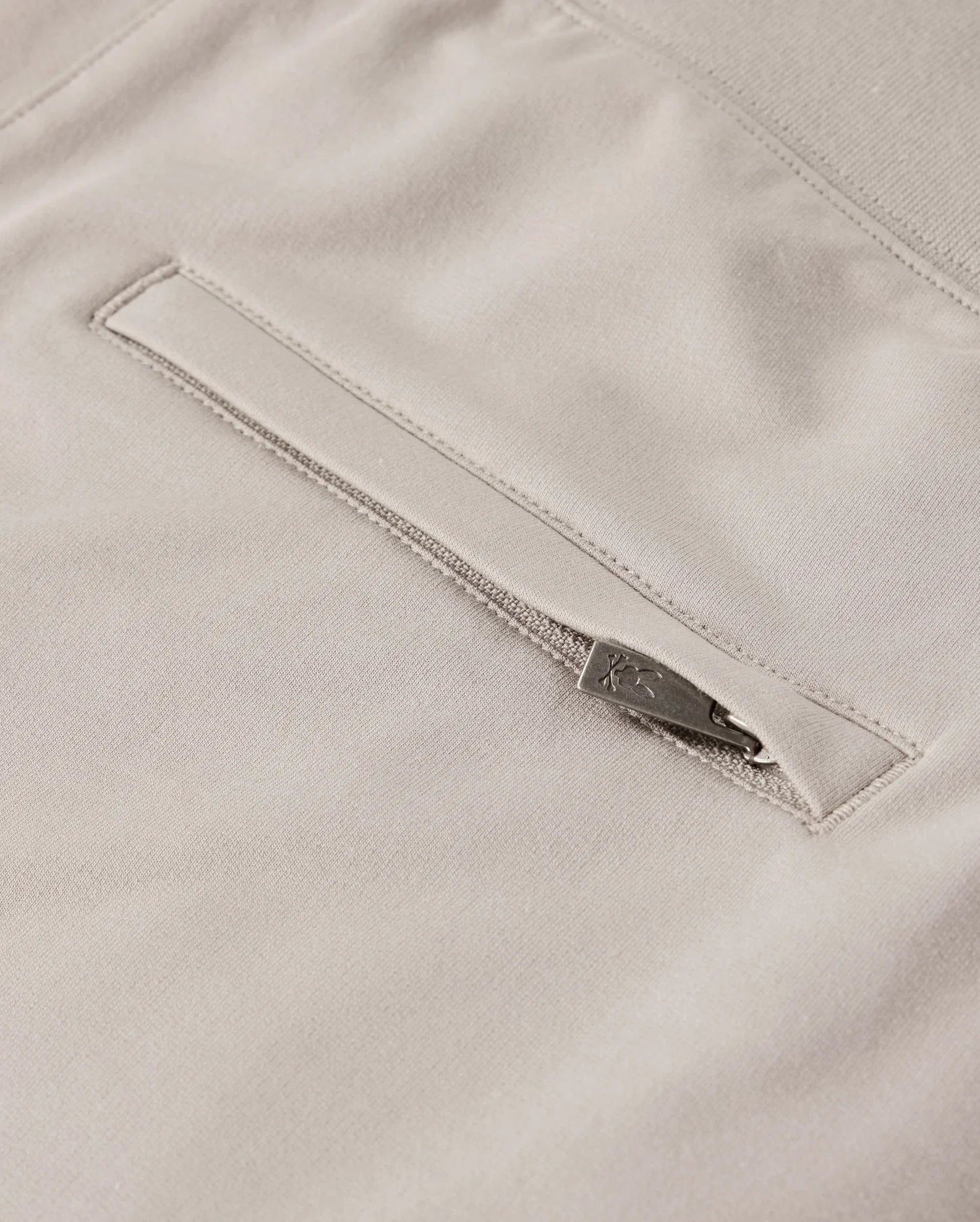 Close-up of the *MENS WALTER LIGHTWEIGHT SWEATSHORT - B6R477D200* by Psycho Bunny showcasing a light beige fabric with a horizontal zippered pocket. The zipper includes a metallic pull tab for easy access, and the surrounding fabric appears soft and smooth. This high-quality cotton blend ensures a uniform, finely woven texture in this lightweight sweatshort.