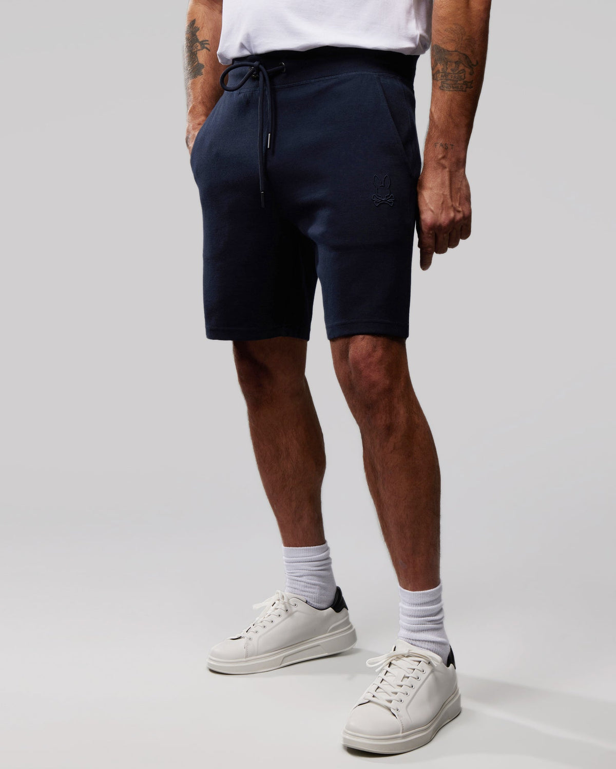 A person wearing a white t-shirt and navy blue Psycho Bunny MENS OUTLINE SWEATSHORTS - B6R507ARCN with an embroidered logo stands against a plain grey background. Their hands are in their pockets, revealing white crew socks and sneakers, while several tattoos peek out from beneath the hems of the MENS OUTLINE SWEATSHORTS - B6R507ARCN.