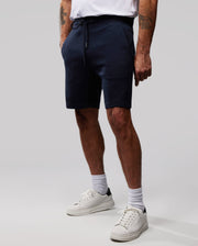A person wearing a white t-shirt and navy blue Psycho Bunny MENS OUTLINE SWEATSHORTS - B6R507ARCN with an embroidered logo stands against a plain grey background. Their hands are in their pockets, revealing white crew socks and sneakers, while several tattoos peek out from beneath the hems of the MENS OUTLINE SWEATSHORTS - B6R507ARCN.
