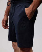 A person is wearing black MENS OUTLINE SWEATSHORTS - B6R507ARCN with a hand in their pocket. The sweatshorts, part of the OUTLINE collection by Psycho Bunny, feature a Psycho Bunny logo with crossbones beneath its head on the left thigh. The person's left forearm has a tattoo that reads "FAST." The background is plain and minimal.
