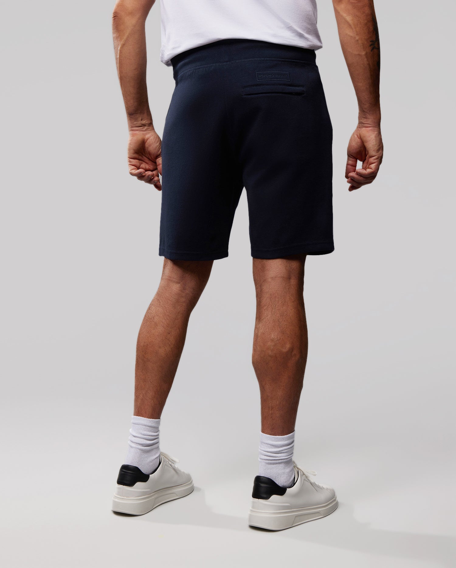 A person stands facing away, wearing a white Psycho Bunny T-shirt, dark navy Psycho Bunny MENS OUTLINE SWEATSHORTS - B6R507ARCN, white socks, and white sneakers with black accents. The background is plain and light gray.
