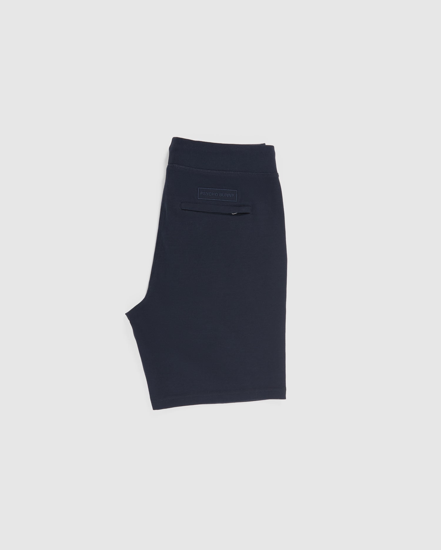 Image of a folded pair of navy blue sweat shorts from the Psycho Bunny collection. The MENS OUTLINE SWEATSHORTS - B6R507ARCN feature a back pocket with a rectangular label above it, displaying some text. The background is plain white.
