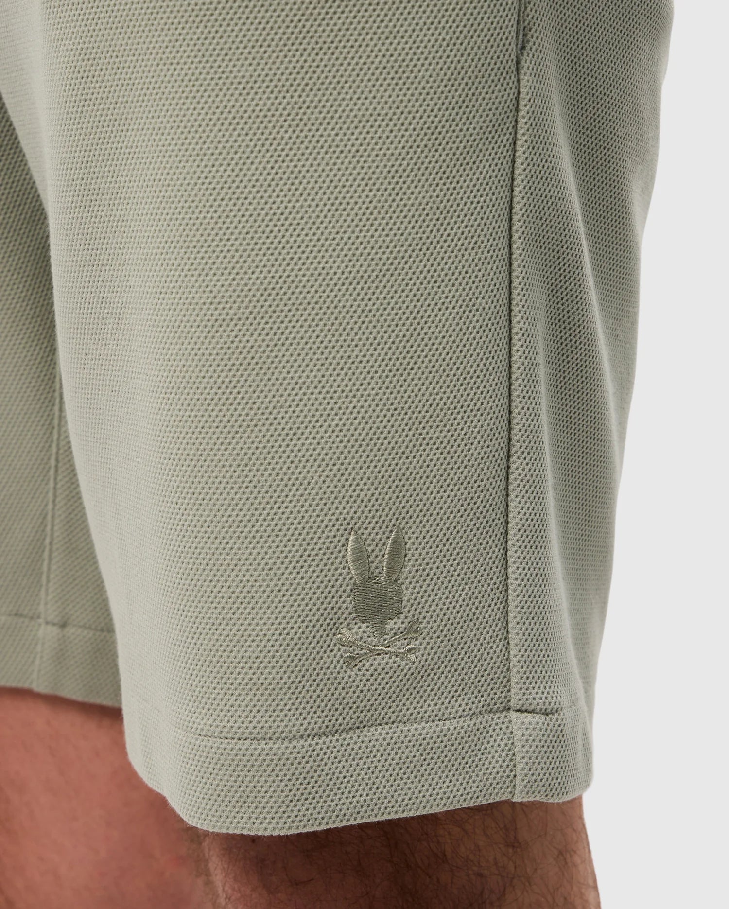 Close-up of a light green, sweat-wicking fabric with a small, white embroidered logo of a rabbit on the lower side near a seam on the Psycho Bunny MENS SHIRO KNIT HONEYCOMB SHORT - B6R567C200.