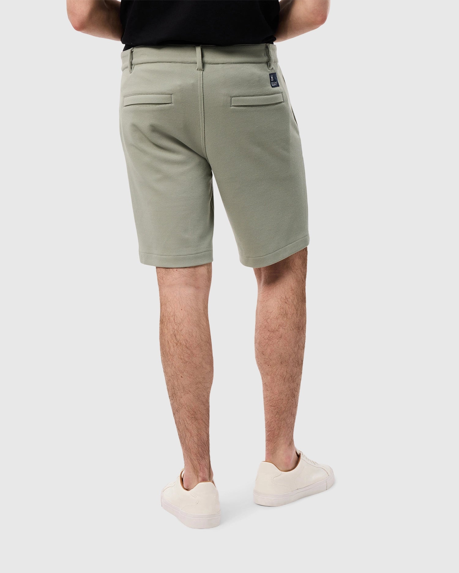 A man wearing Psycho Bunny sage green knit shorts and white sneakers stands facing away from the camera, showing the back design of the MENS SHIRO KNIT HONEYCOMB SHORT - B6R567C200 with a small label on the right pocket.