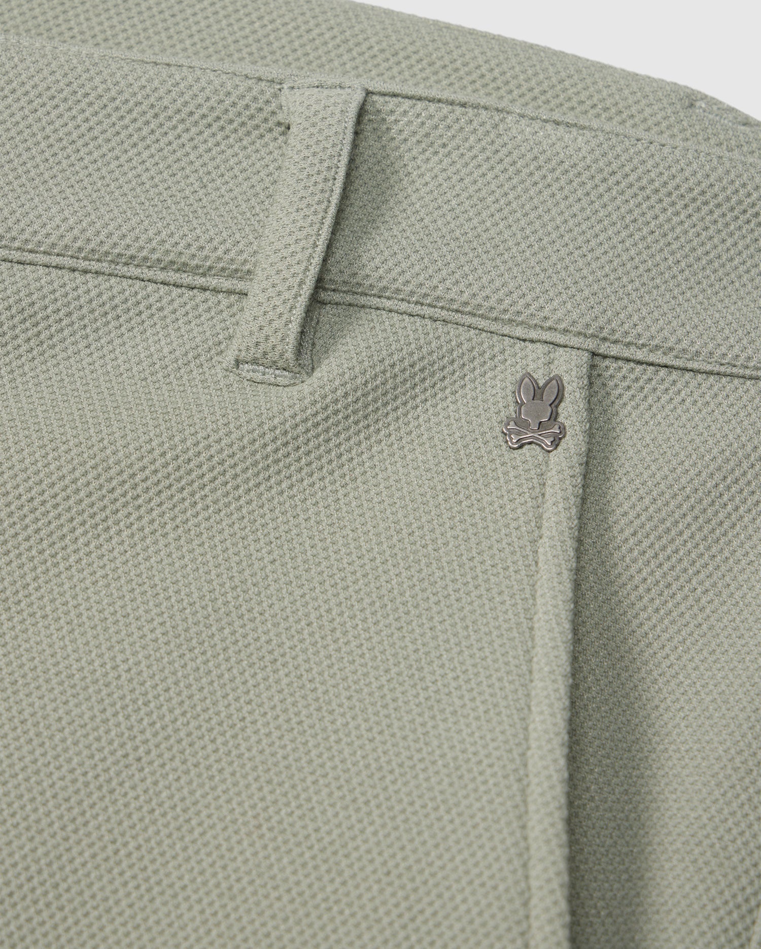 Close-up of a light green Psycho Bunny honeycomb fabric with a subtle texture, featuring a small metal bunny logo near the seam of a pocket.