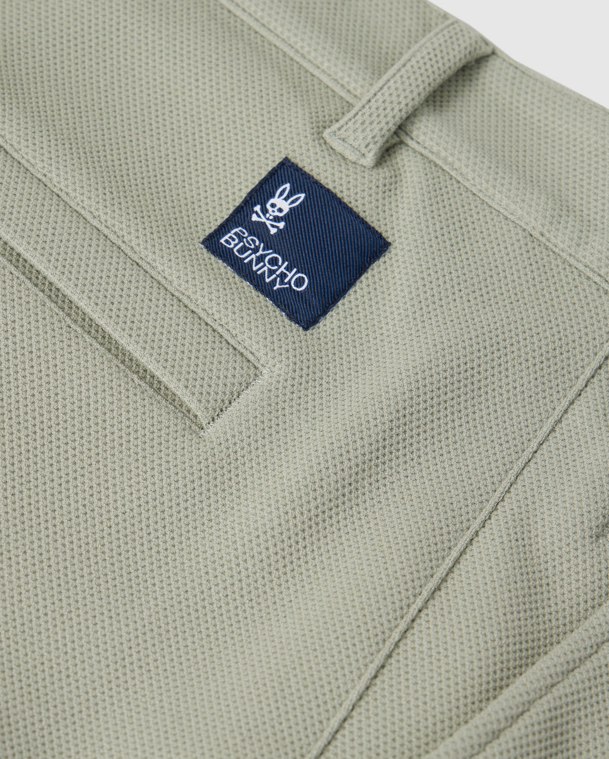 Close-up of a green Psycho Bunny honeycomb fabric material featuring a stitched pocket detailing and a small blue label with the logo "Psycho Bunny" on the upper part of the pocket.