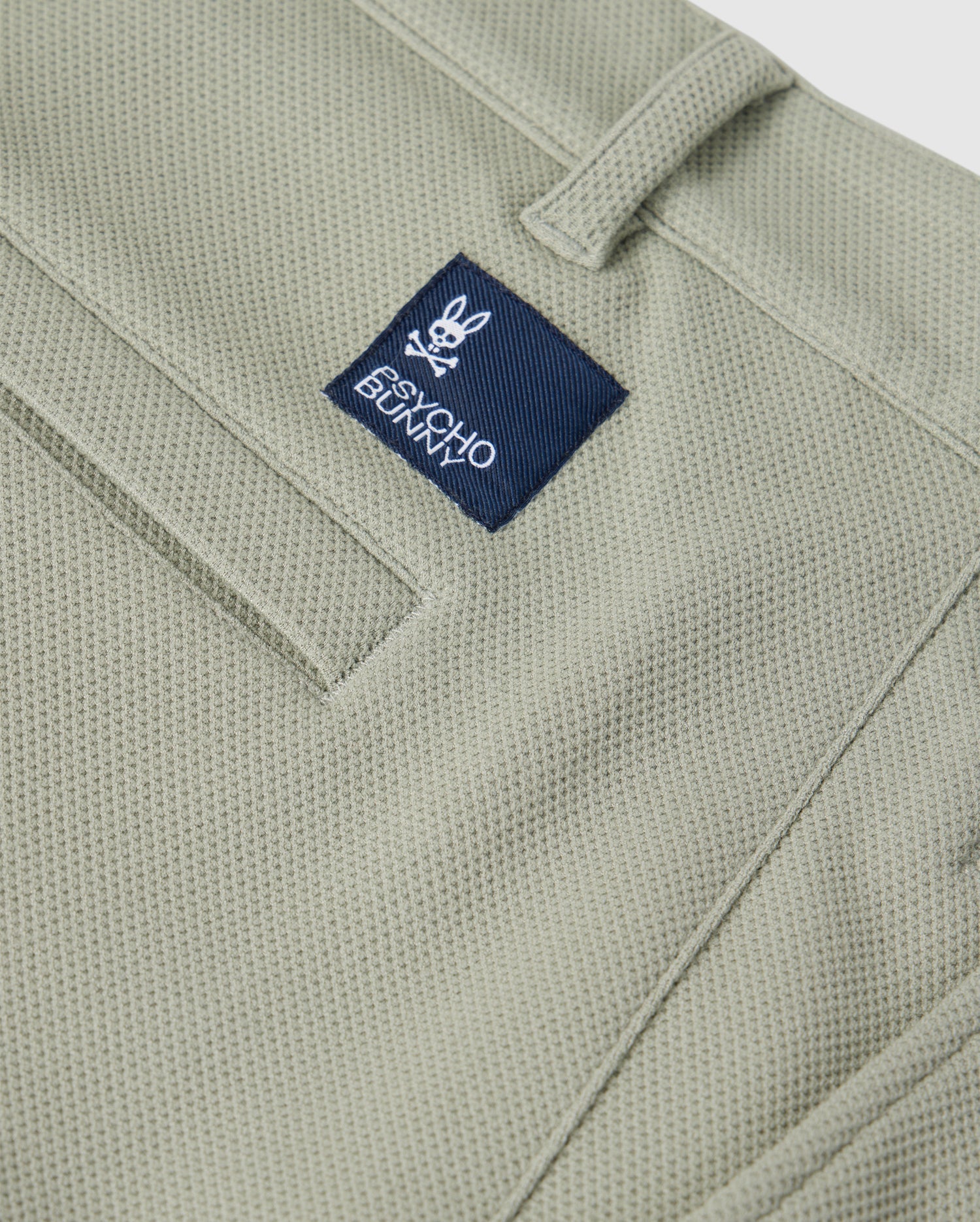 Close-up of a green Psycho Bunny honeycomb fabric material featuring a stitched pocket detailing and a small blue label with the logo "Psycho Bunny" on the upper part of the pocket.