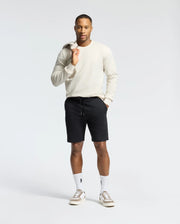 A man exudes confidence in a light sweater and Psycho Bunny's Levanzo Embroidered Micro French Terry Sweatshorts (B6R578F200). With one hand on his sweater collar, complemented by white socks and tennis shoes, the micro French terry fabric merges seamlessly with the plain white backdrop.