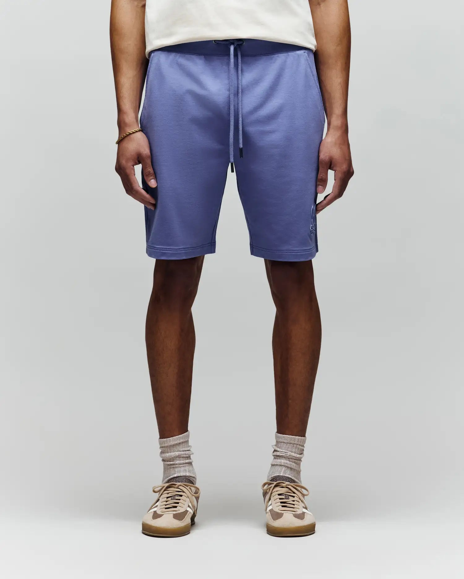 A person stands facing forward, wearing Psycho Bunny MENS LEVANZO FRENCH TERRY SWEATSHORT - B6R578F200 in light purple, paired with a white shirt, beige socks, and beige sneakers against a plain light background. Image is cropped from shoulders down.