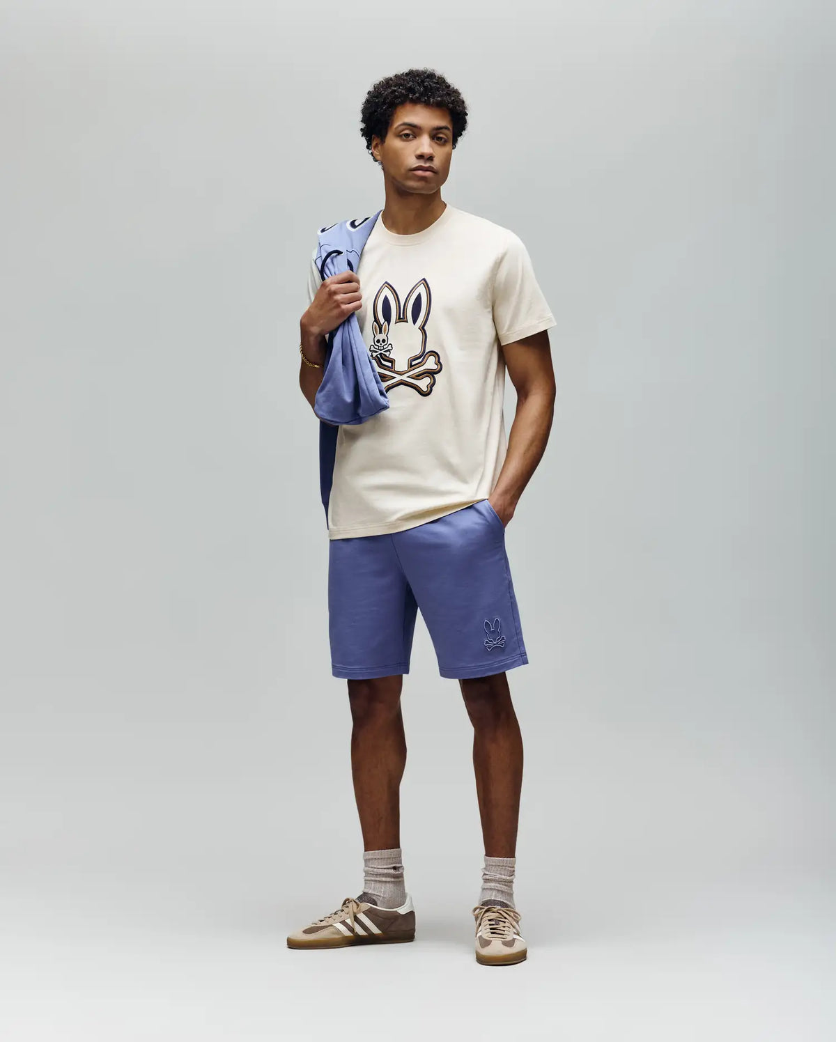 A man stands against a plain backdrop wearing a cream T-shirt with a bunny graphic, Psycho Bunny MENS LEVANZO FRENCH TERRY SWEATSHORT - B6R578F200 in light purple, beige socks, brown sneakers, and holds a matching sweatshirt over his shoulder.