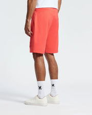 A person is wearing Psycho Bunny's MENS LEVANZO FRENCH TERRY SWEATSHORT - B6R578F200 in red, paired with a white t-shirt and white socks featuring a black logo, highlighting their comfort against a plain, light background while sporting white sneakers.