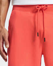 Someone is wearing Psycho Bunny's coral MENS LEVANZO FRENCH TERRY SWEATSHORT with a drawstring waistband, a relaxing fit with their left hand at their side, paired with a white shirt.