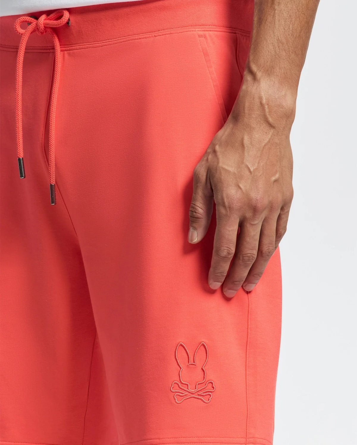 A person's hand rests on the comfortable Psycho Bunny MENS LEVANZO FRENCH TERRY SWEATSHORT in coral. These shorts, adorned with a rabbit skull and crossbones logo, feature a drawstring with metal tips, against a plain white background.