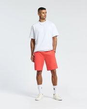 A man is wearing a white t-shirt, Psycho Bunny's Levanzo French Terry Sweatshorts in red with a subtle logo on the left, and white socks and sneakers. Made from micro French terry for comfort, he gazes slightly right against a plain white backdrop.