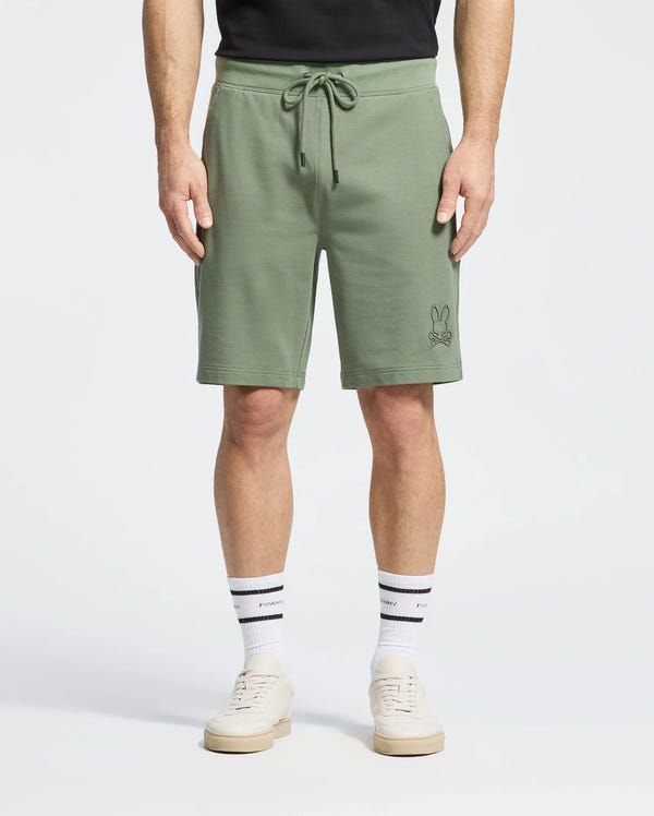 Man wears Psycho Bunny's olive green Levanzo French Terry Sweatshorts B6R578F200 with a small embroidered Bunny near the left pocket, featuring a drawstring waist. He pairs them with a black shirt, white sports socks, and beige sneakers.