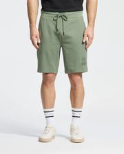 Man wears Psycho Bunny's olive green Levanzo French Terry Sweatshorts B6R578F200 with a small embroidered Bunny near the left pocket, featuring a drawstring waist. He pairs them with a black shirt, white sports socks, and beige sneakers.