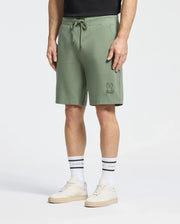 A person is wearing green Psycho Bunny Levanzo French Terry Sweatshorts with an embroidered Bunny design on the left leg, white socks with black text, beige sneakers, and a black shirt against a plain white background.