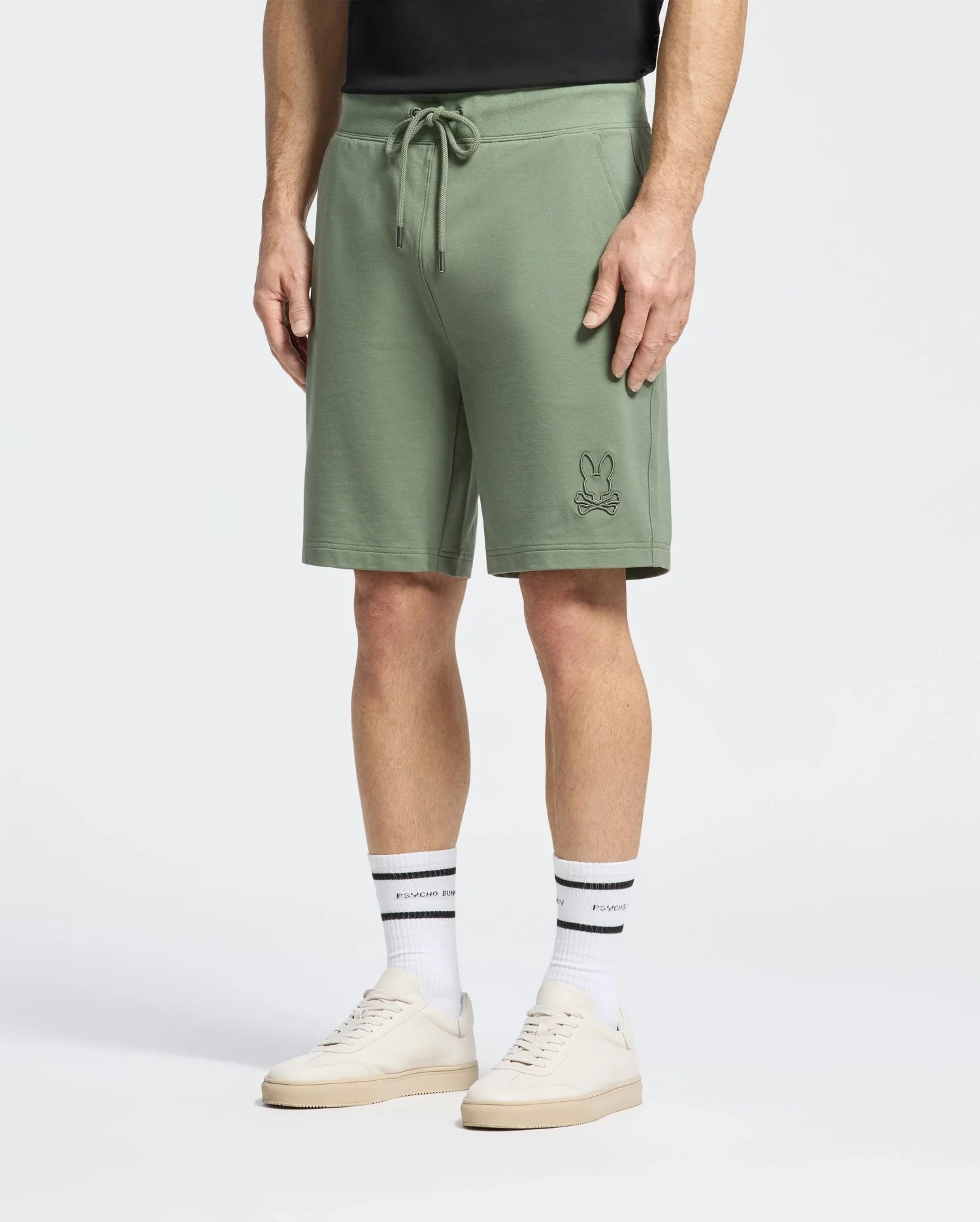 A person is wearing green Psycho Bunny Levanzo French Terry Sweatshorts with an embroidered Bunny design on the left leg, white socks with black text, beige sneakers, and a black shirt against a plain white background.