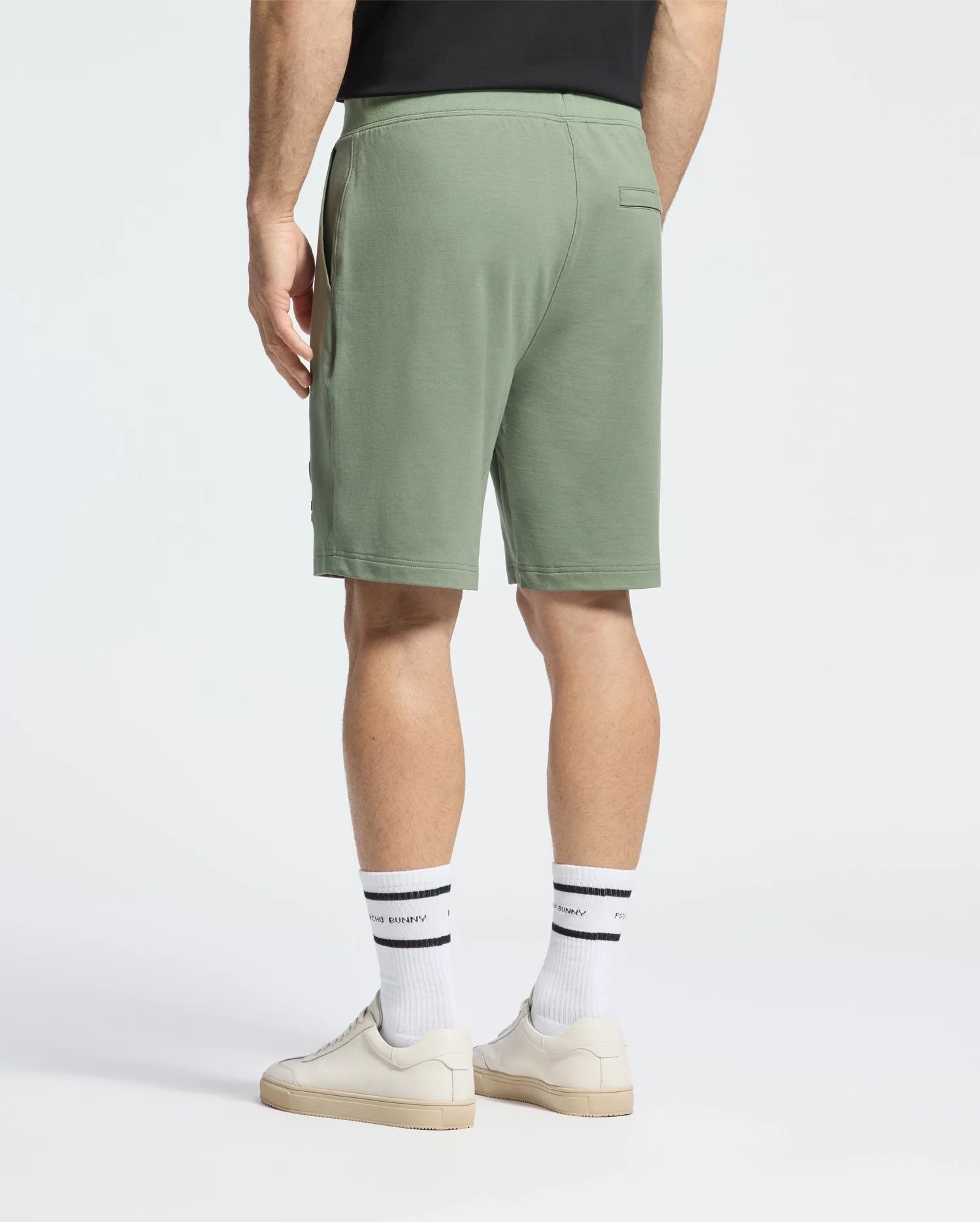 A person is standing with their back to the camera against a white background, wearing green Psycho Bunny Levanzo French Terry Sweatshorts, a black shirt, white socks with black stripes and lettering, and white sneakers.