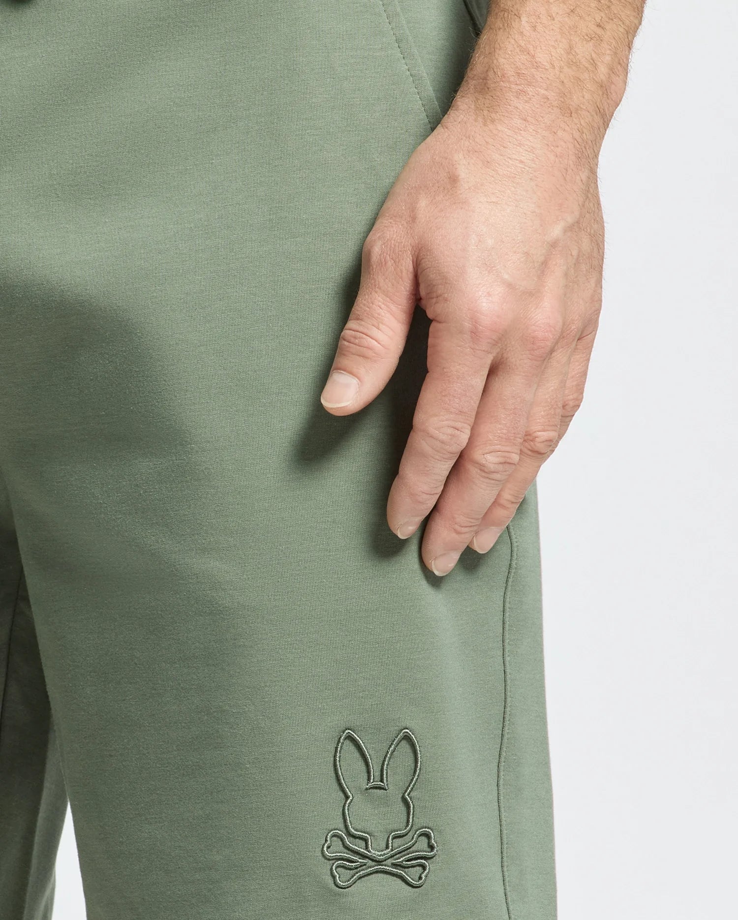 A close-up showcases a hand on olive green pants made from micro French terry, featuring an embroidered Bunny and crossbones logo by the pocket. Set against a plain white background, it exudes the sporty elegance of Psycho Bunny's Levanzo Sweatshorts (B6R578F200).