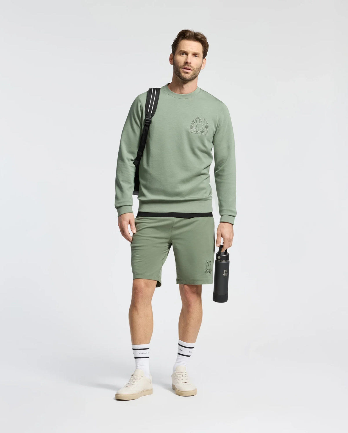 A man stands against a plain backdrop in a matching light green micro French terry sweatshirt and MENS LEVANZO FRENCH TERRY SWEATSHORT, holding a black Psycho Bunny water bottle and sporting a black backpack. His look is completed with white sneakers and striped socks.