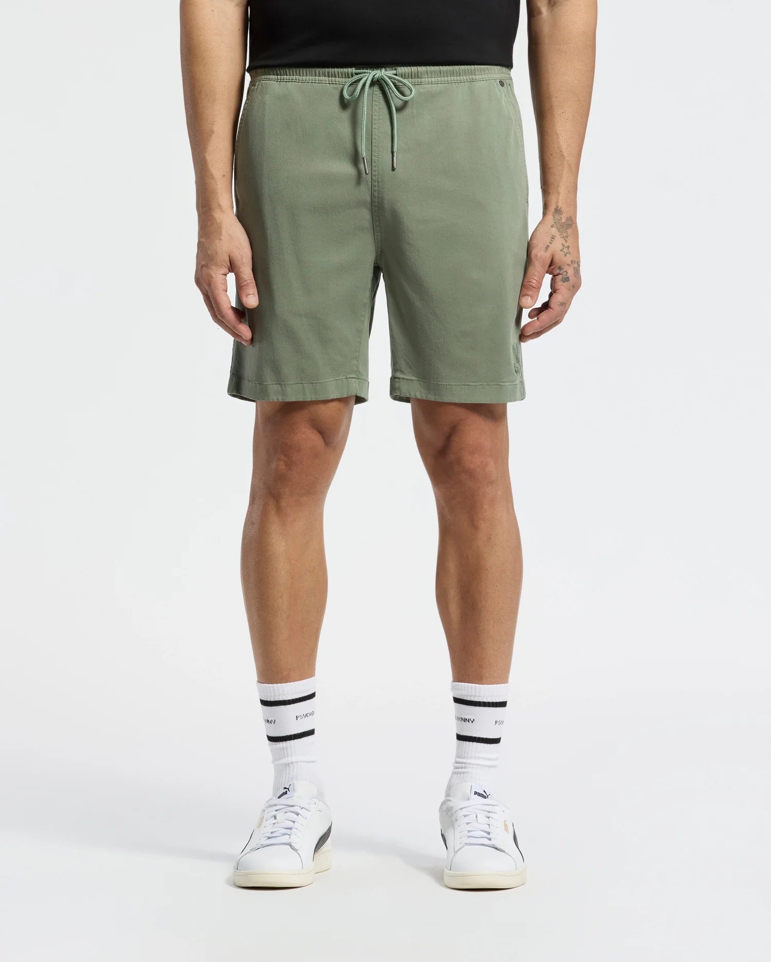 A person stands wearing light green MENS WILLIS STRETCH TENCEL SHORTS by Psycho Bunny with an embroidered Bunny Drawstring, a partially visible black shirt, white crew socks with black stripes, and white sneakers. Their arms are relaxed by their sides, showcasing effortless style and a tattoo on one hand.