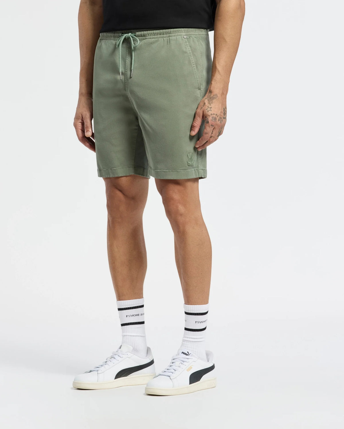 Wearing olive green MENS WILLIS STRETCH TENCEL SHORT - B6R584F200 by Psycho Bunny and a black shirt, paired with white striped socks, they don sleek white sneakers with a black logo. Their tattoos add flair to the plain white backdrop.