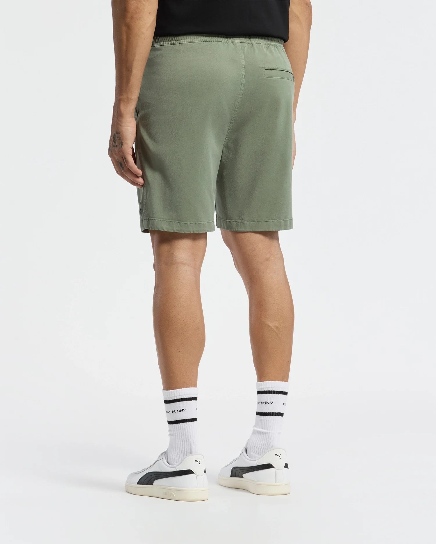 A person effortlessly showcases style while wearing Psycho Bunny's MENS WILLIS STRETCH TENCEL SHORT - B6R584F200 in green with a back pocket, paired with black-stripe white socks. Their look is completed by a black shirt and embroidered Bunny Drawstring sneakers, all set against a plain white backdrop.