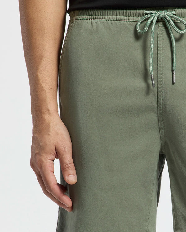 A person effortlessly exudes style in Psycho Bunny's Willis Stretch Tencel Shorts, olive green with a drawstring waist, as their left hand rests by their side against a plain white backdrop.