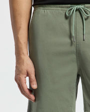 A person effortlessly exudes style in Psycho Bunny's Willis Stretch Tencel Shorts, olive green with a drawstring waist, as their left hand rests by their side against a plain white backdrop.
