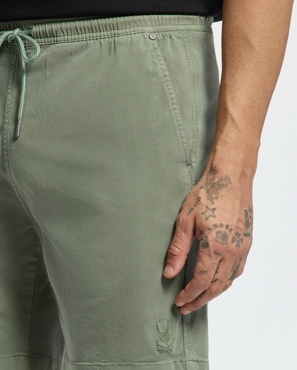 A close-up shows a person in Psycho Bunny's Men’s Willis Stretch Tencel Shorts in light green, featuring an embroidered bunny on the drawstring. The tattooed right hand rests alongside, against a plain white background.