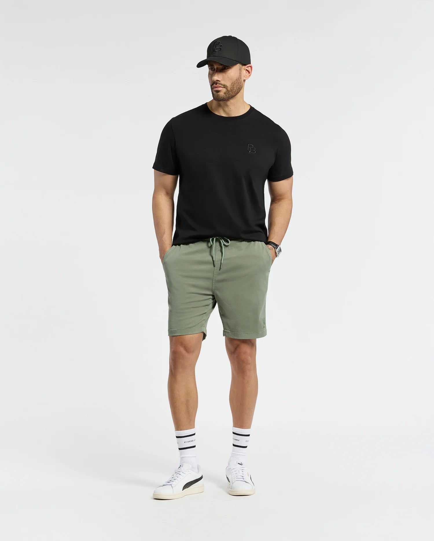 A man embodies effortless style in a black cap, t-shirt, and Psycho Bunny's MENS WILLIS STRETCH TENCEL SHORT in green. White sneakers paired with striped socks add a classic touch to his minimalistic look, set against a plain white backdrop where his understated confidence speaks volumes.