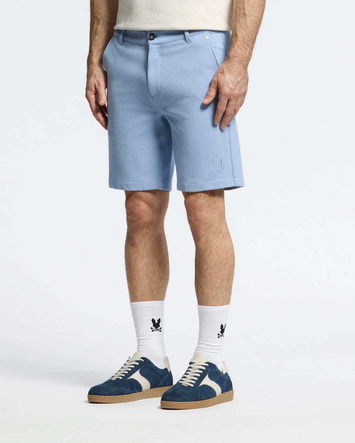 A person wearing the light blue Psycho Bunny MENS SHIRO KNIT HONEYCOMB SHORT – B6R585F200, paired with logoed white socks. Blue and white sneakers add flair, while an embroidered bunny subtly adorns the shorts. The focus remains on their lower body against a plain background.