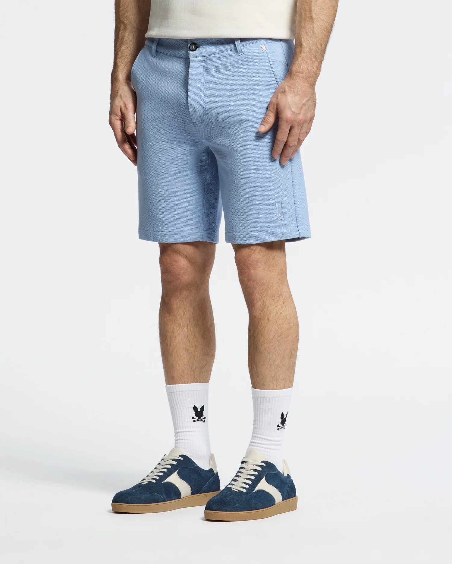 A person wearing the light blue Psycho Bunny MENS SHIRO KNIT HONEYCOMB SHORT – B6R585F200, paired with logoed white socks. Blue and white sneakers add flair, while an embroidered bunny subtly adorns the shorts. The focus remains on their lower body against a plain background.