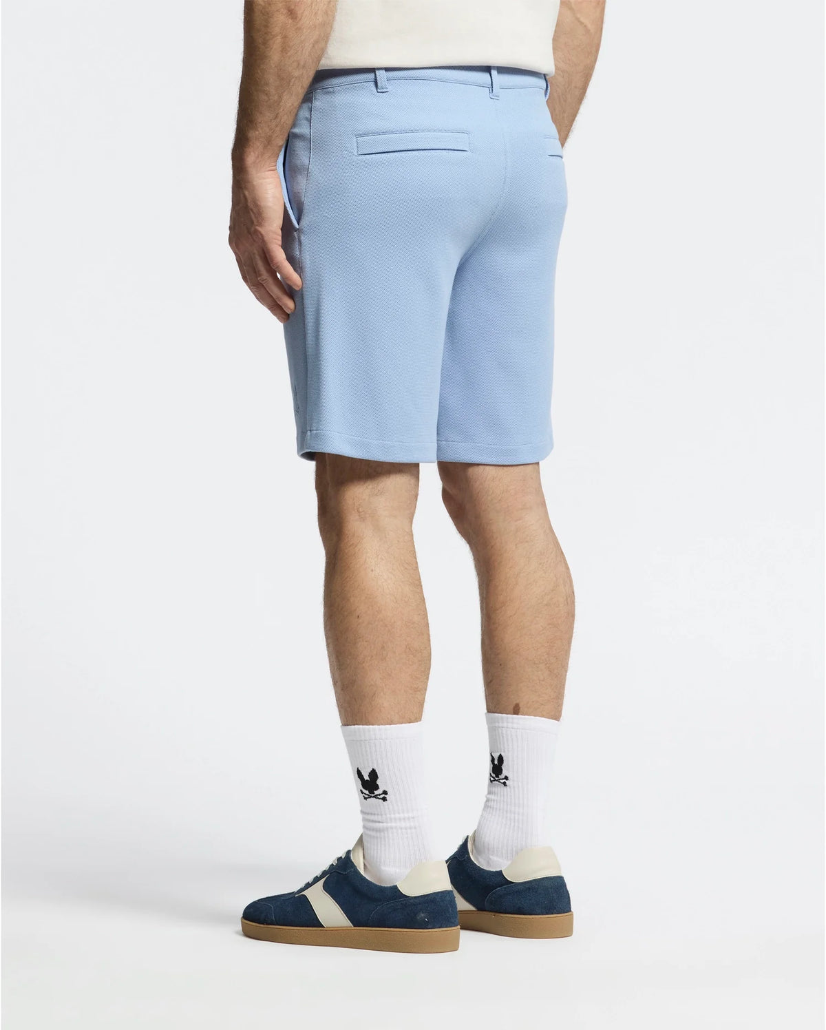 Someone wearing Psycho Bunny's MENS SHIRO KNIT HONEYCOMB SHORT in light blue stands facing away, showcasing white socks with an embroidered bunny and navy blue sneakers with white accents. The plain, light background highlights the outfit's details.