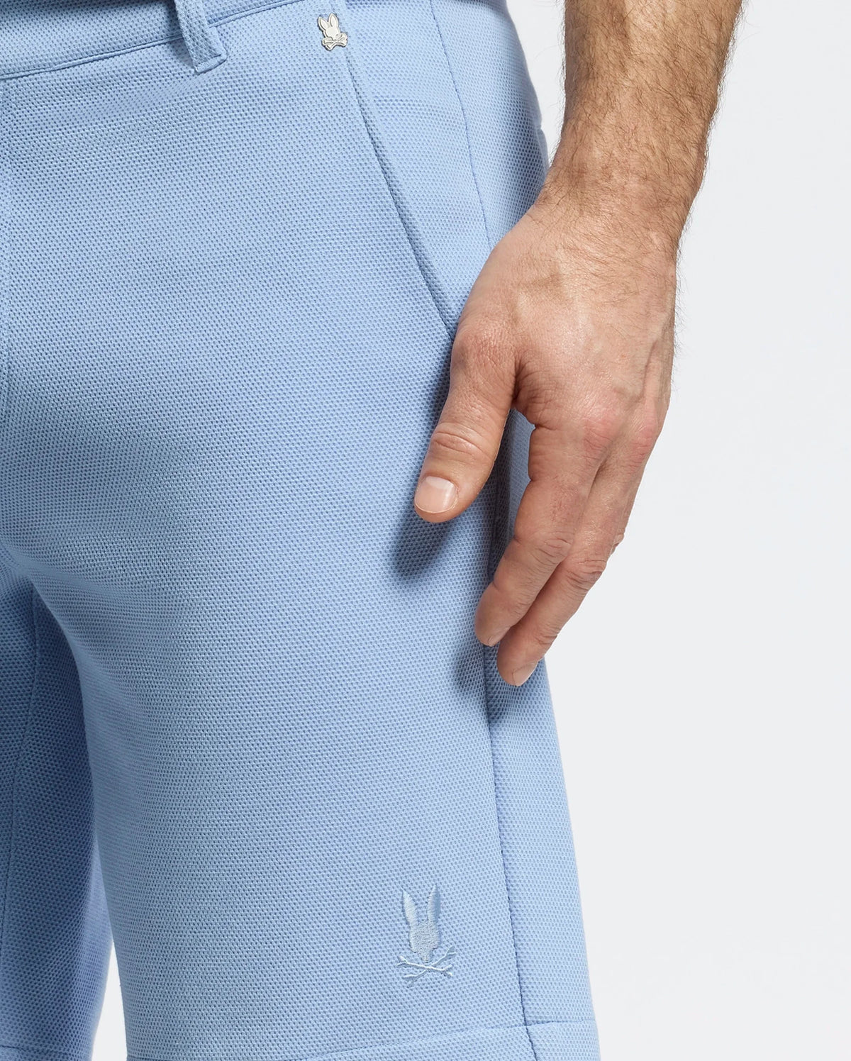 A person models Psycho Bunny's MENS SHIRO KNIT HONEYCOMB SHORT (B6R585F200) in light blue with a honeycomb pattern and small embroidered logo near the hem, resting a hand by the versatile shorts against a plain white background.
